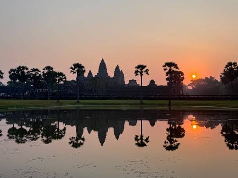       Iconic sunrise view of Angkor Wat reflected perfectly in a still pond with palm trees silhouetted.
  