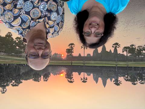       Smiling couple taking a selfie in front of Angkor Wat at sunrise with its reflection in the pond.
  