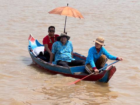       Three people in a narrow blue boat paddling across a wide muddy river under bright light.
  