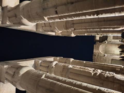       Night-time view between massive lit columns inside an Egyptian temple, with visitors silhouetted at the base.
  