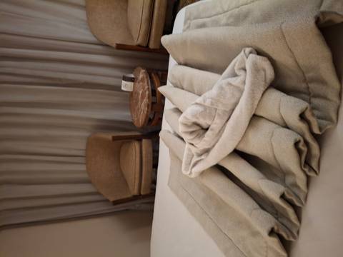       Hotel or cruise-ship cabin with folded beige towels on the bed, two chairs and a small round table in soft lighting.
  