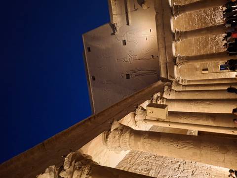       Night view of a hieroglyph-covered temple courtyard with lit columns against a deep blue sky, crowds below.
  
