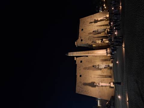       Lit statues and obelisk at Luxor Temple courtyard at night with many visitors gathered below.
  