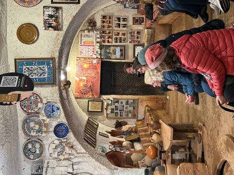       Visitors seated inside a pottery studio adorned with decorated plates and photos as a host demonstrates craft.
  