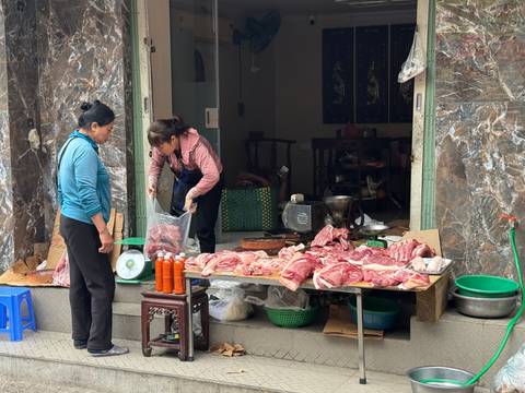       Street-side butcher shop with slabs of fresh meat laid out on boards and shoppers conversing.
  