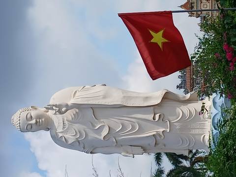       Tall white Buddha statue making a gesture beside the Vietnamese national flag waving against blue sky.
  