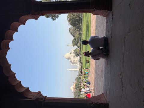       Couple sitting under an arched pavilion gazing at the distant Taj Mahal across manicured lawns.
  