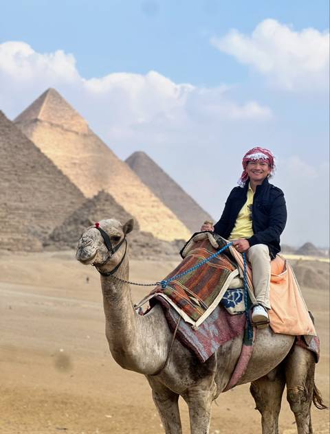       Traveller riding a camel with the Great Pyramids of Giza rising majestically behind.
  