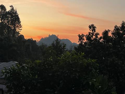       Soft orange and pink hues paint the sky behind silhouetted hills and treetops at dusk.
  