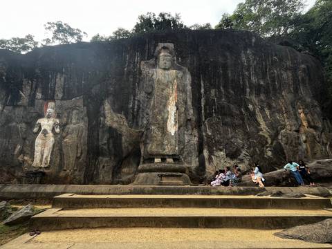       Ancient rock cliff with large standing Buddha statues carved into the stone, several visitors sitting at the base.
  
