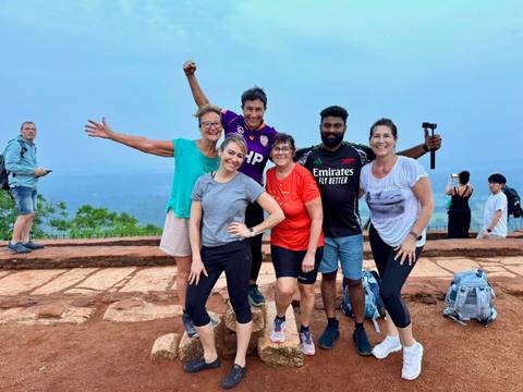       Smiling travellers pose on a brick terrace with Sigiriya rock looming in the background.
  