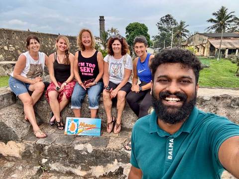       Group relaxes on a stone wall near coastal ruins holding a holiday sign.
  