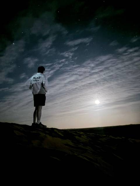       Person standing on a ridge at night looking out at a bright moon and streaked clouds.
  