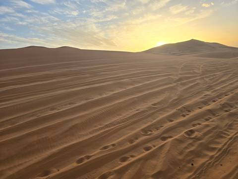       Rippled tracks across soft sand dunes with the sun setting behind a ridge, casting warm light.
  