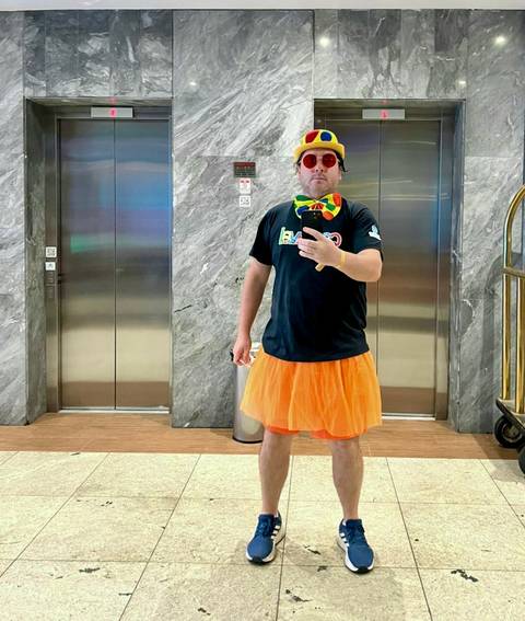       Man taking a mirror selfie wearing an orange tutu and colorful bow tie in a hotel lobby.
  