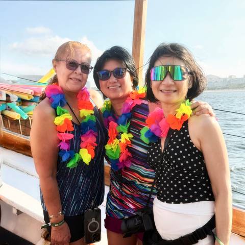       Three women wearing colorful leis pose on a boat with coastal cityscape behind.
  