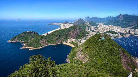       Stunning aerial view of Rio de Janeiro’s coastline, Sugarloaf Mountain, and cityscape against a bright blue sky and sea.
  