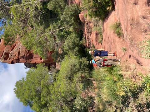       Small hiking party walks along red sandstone trail beneath towering cliff and pine trees in Catalonia
  