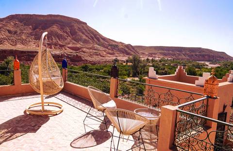       Scenic terrace of a riad with hanging chair overlooking red cliffs and oasis below
  