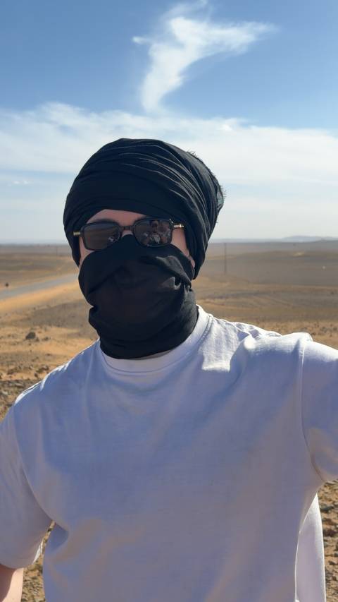       Close-up portrait of a traveler wearing sunglasses and a desert head scarf with sandy plains behind.
  