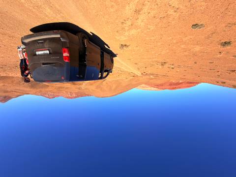       A dusty van parked in a vast arid landscape of red and tan hills under clear skies.
  