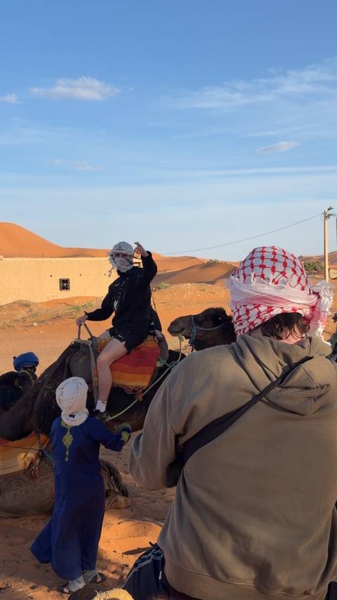       Travelers riding camels across orange desert dunes, one rider gesturing forward.
  