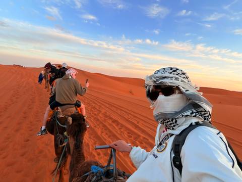       A line of camel riders traverses bright orange dunes at sunset, lead rider in foreground wearing sunglasses and scarf.
  