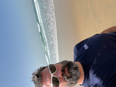       Man taking selfie on a sunny sandy beach with breaking waves behind
  