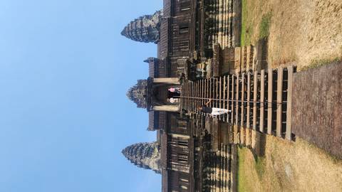       Visitors climb steep wooden stairs to the towering central sanctuary of Angkor Wat under a clear blue sky.
  