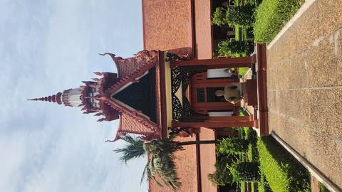       Ornate Khmer-style pavilion with red tiled roof and a seated Buddha statue in a manicured courtyard under a bright blue sky.
  