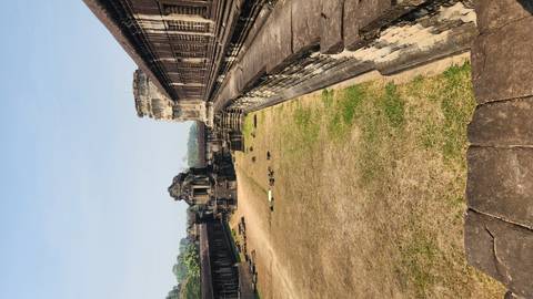       Long sandstone galleries and central courtyard of Angkor Wat ruins under clear skies, showing weathered carvings and open grass.
  