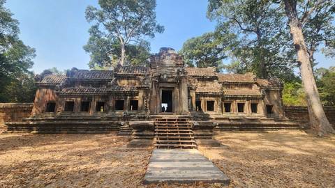       Stone temple ruin nestled among tall trees with a central staircase leading to multiple windowed chambers, carpeted by dry leaves.
  