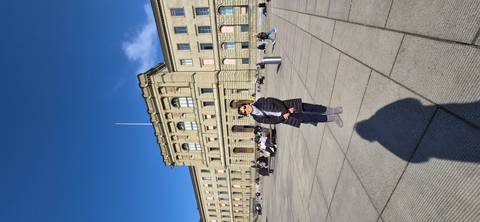       Visitor poses in a spacious plaza before the stately sandstone main building of ETH Zürich under a cloudless sky.
  