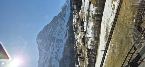       Broad valley floor with patchy snow and railway, dominated by a towering alpine peak against a clear sky.
  