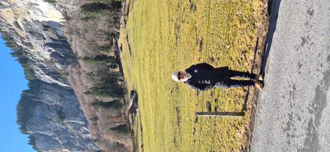       Visitor in winter coat stands beside a pasture backed by steep rocky cliffs and mixed conifer forest.
  