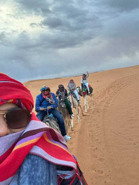       A line of tourists riding camels across rolling orange dunes, with the lead rider snapping a colourful scarf-covered selfie.
  