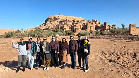       A tour group stands on dusty ground with the earthen citadel of Aït Benhaddou rising behind them.
  