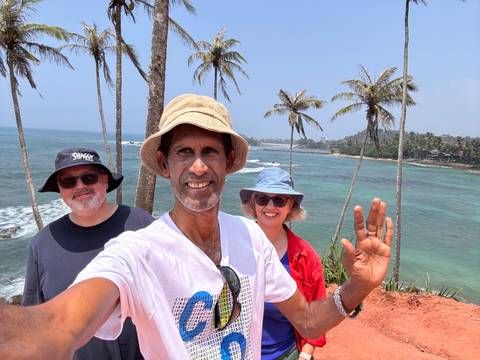       Smiling trio taking a selfie on a coastal cliff lined with palm trees and turquoise sea in the background.
  