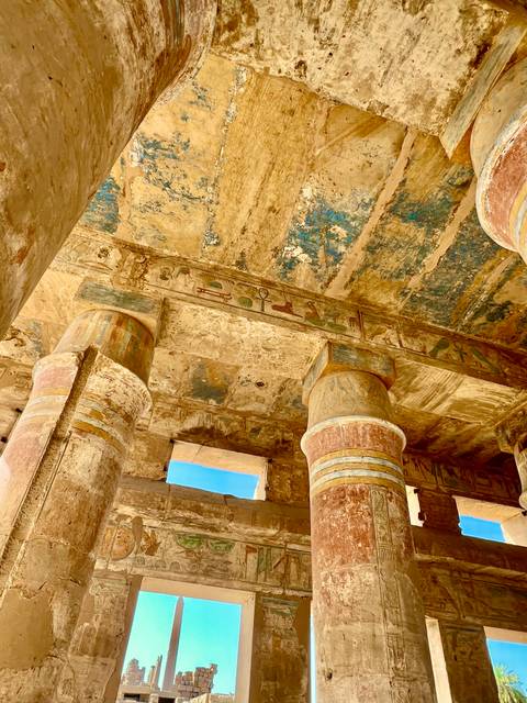       Weathered stone columns and a richly painted ceiling displaying ancient Egyptian art bathed in warm light.
  