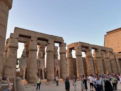       Crowds gather among colossal statues and sandstone columns at Luxor Temple in the soft evening light.
  