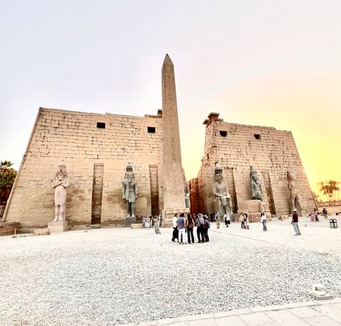       Obelisk and massive pylons of Luxor Temple glow against a colorful sunset sky while visitors explore the forecourt.
  