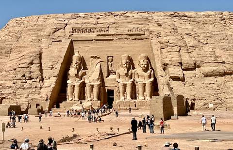       Grand rock-cut façade of Abu Simbel with four colossal seated statues overlooking a sandy forecourt filled with tourists.
  