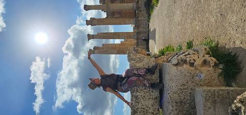       Traveler playfully posing on ancient stone at Jerash archaeological site with towering columns and bright midday sun overhead.
  