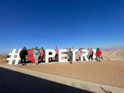       Tour group posing in front of giant #❤️PETRA sign under a clear blue sky overlooking the desert hills.
  