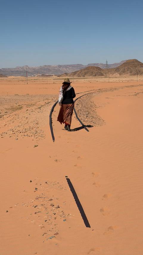       Woman walking along an abandoned desert rail track, footprints trailing behind in the orange sand of Wadi Rum.
  