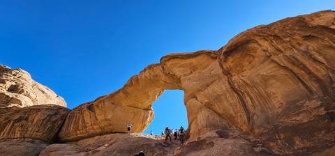       Spectacular sandstone arch in Wadi Rum with adventurers standing triumphantly on top against a vivid blue sky.
  