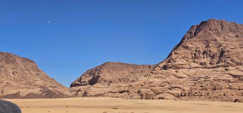       Wide desert valley framed by rugged sandstone mountains under a cloudless sky with the moon visible in daylight.
  