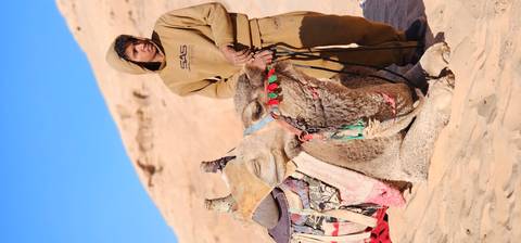       Bedouin camel handler in tan clothing standing beside a resting camel adorned with colorful reins in Wadi Rum.
  