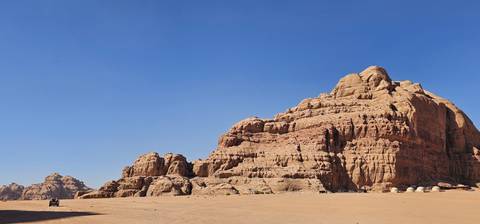       Expansive desert landscape with towering red sandstone massif under a clear blue sky and faint vehicle tracks in the sand.
  