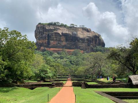       Iconic Sigiriya Rock Fortress rising above dense jungle with the ancient brick entrance path leading toward it.
  